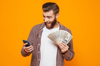 Cep telefonu, para banknot gösterilen tutan sarı arka plan üzerinde izole ayakta rahat kıyafetler giymiş neşeli genç bir adam portresi