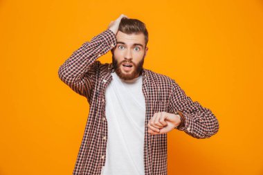 Akıllı saatini kontrol sarı arka plan üzerinde izole ayakta rahat kıyafetler giymiş şok genç bir adam portresi