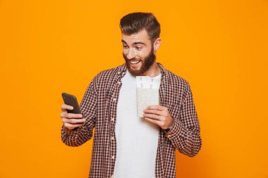 Cep telefonu kullaranarak pasaport, holding sarı arka plan üzerinde izole ayakta rahat kıyafetler giymiş neşeli genç bir adam portresi
