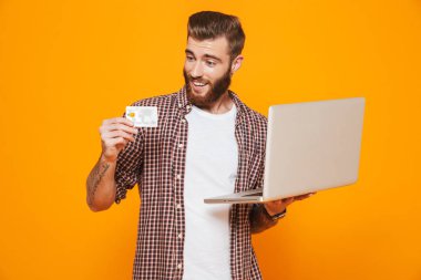 Dizüstü bilgisayar, plastik kredi kartı gösteren holding sarı arka plan üzerinde izole ayakta rahat kıyafetler giymiş neşeli genç bir adam portresi