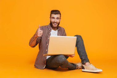 Dizüstü bilgisayar tutan sarı arka plan üzerinde izole oturma rahat kıyafetler giymiş neşeli genç bir adam portresi