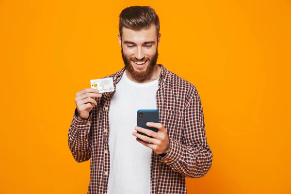 一个开朗的年轻人穿着休闲服站在黄色背景上 拿着手机 展示塑料信用卡的肖像 — 图库照片