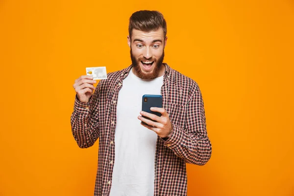 一个开朗的年轻人穿着休闲服站在黄色背景上 拿着手机 展示塑料信用卡的肖像 — 图库照片