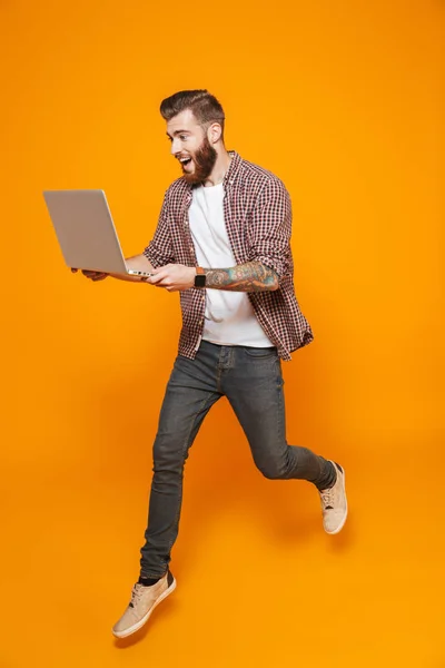 一个开朗的年轻人穿着在黄色背景中被隔离的休闲服 拿着笔记本电脑 跳来跳的全长画像 — 图库照片