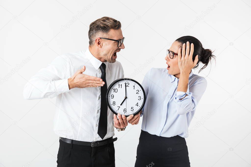 Couple of shocked colleagues wearing formal clothing standing isolated over white background, showing wall clock
