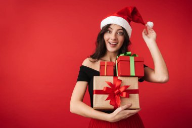 Hediye kutuları yığını tutan kırmızı arka plan üzerinde Noel şapka ayakta giyen heyecanlı genç kadın izole