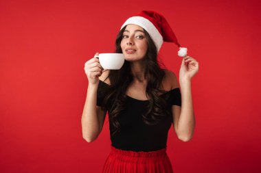 Sıcak çikolata tutan kırmızı arka plan üzerinde duran Noel şapka neşeli genç kadın izole
