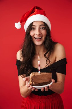 Fotoğraf güzel bir genç kadının kek Noel şapka tutan kırmızı duvar arka plan üzerinde izole ayakta.