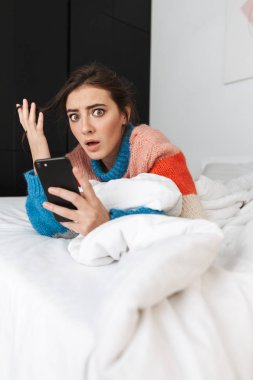 Evde yatakta yatan ve cep telefonu kullanarak hoşnutsuz kadın 20'li yaşlarda giyen kazak fotoğrafı