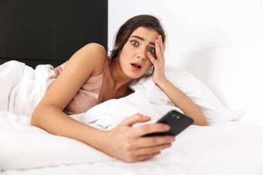 Heyecanlı kadın 20'li yaşlarda koyu saçlı smartphone beyaz yastık üzerinde yatakta uyku veya nap sonra yalan Ise tutarak fotoğrafı