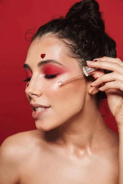Bella giovane donna con trucco rosso luminoso isolato su sfondo muro rosso in posa con cuori sul viso tenuta siero idratante . — Foto Stock