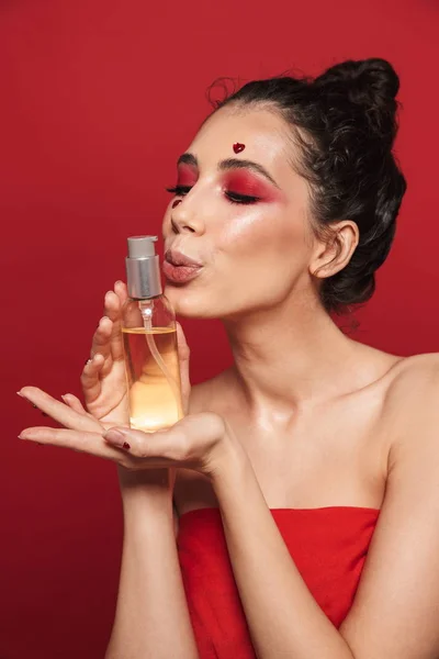 Smuk ung kvinde med rød lys makeup isoleret over rød væg baggrund udgør med hjerter på ansigt holder olie . - Stock-foto