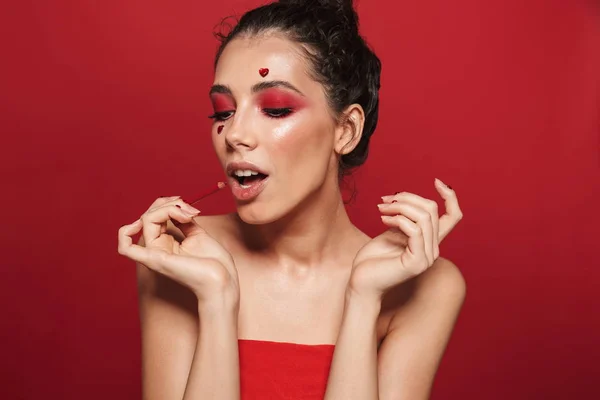 Όμορφη νεαρή γυναίκα με κόκκινο λαμπερό μακιγιάζ που απομονώνονται σε φόντο κόκκινο τοίχο ποζάρει με καρδιές στο πρόσωπο κρατά μολύβι χειλιών. — Φωτογραφία Αρχείου