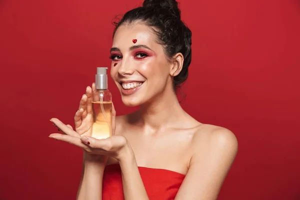 Smuk ung kvinde med rød lys makeup isoleret over rød væg baggrund udgør med hjerter på ansigt holder olie . - Stock-foto