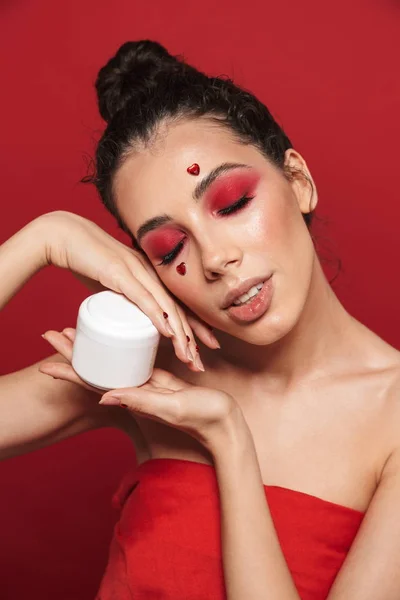 Schöne junge Frau mit rotem hellen Make-up isoliert über roter Wand Hintergrund posiert mit Herzen im Gesicht hält Creme. — Stockfoto