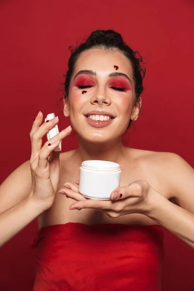 Piękna młoda kobieta z czerwony jasny makijaż na białym tle czerwone ściany pozowanie z sercami na twarzy trzyma krem. — Zdjęcie stockowe