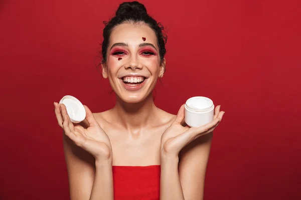 Hermosa mujer joven con rojo brillante maquillaje aislado sobre fondo rojo de la pared posando con corazones en la cara celebración de crema . — Foto de Stock