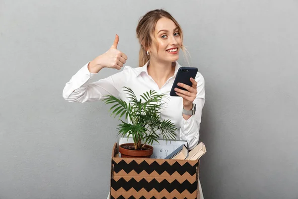 Donna d'affari isolato su sfondo grigio muro che tiene la scatola con pianta e documenti utilizzando il telefono cellulare fare pollici in su gesto . — Foto Stock