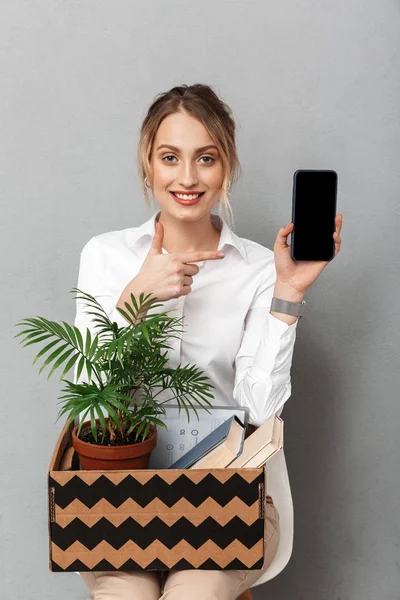 Zakenvrouw geïsoleerd over grijs muur achtergrond houden doos met plant en documenten met behulp van de mobiele telefoon tonen weergeven. — Stockfoto