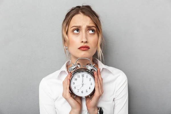 Mujer de negocios confusa aislada sobre fondo de pared gris sosteniendo reloj despertador . —  Fotos de Stock