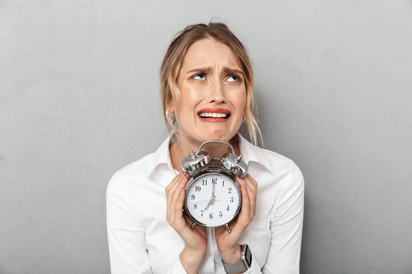 Confused business woman isolated over grey wall background holding alarm clock. — Stock Photo, Image