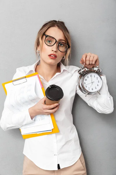 Mujer de negocios confusa aislada sobre fondo de pared gris sosteniendo reloj despertador y café . —  Fotos de Stock