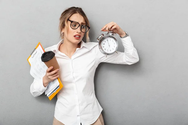 Mujer de negocios confusa aislada sobre fondo de pared gris sosteniendo reloj despertador y café . —  Fotos de Stock