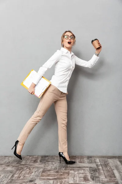 Wanita bisnis terisolasi di atas latar belakang dinding abu-abu yang berjalan sambil memegang kopi dan papan klip . — Stok Foto