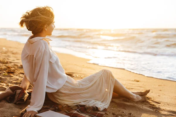 Зображення красивої жінки 20s сидить на піску і дивиться на море, — стокове фото