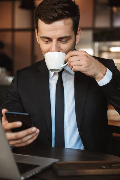 Heureux jeune homme d'affaires assis dans un café en utilisant un ordinateur portable boire du café à l'aide d'un téléphone mobile . — Photo
