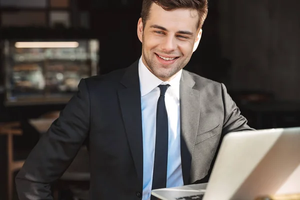 Guapo joven hombre de negocios en ropa formal en el interior de la cafetería con ordenador portátil . — Foto de Stock