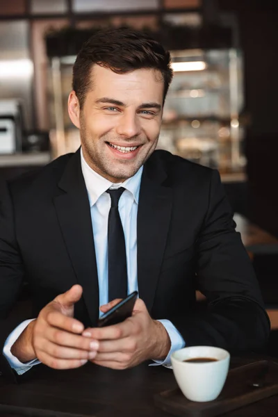 Guapo joven hombre de negocios en ropa formal en el interior de la cafetería con teléfono móvil . — Foto de Stock