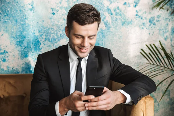 Happy young businessman sitting in cafe using mobile phone. — Stock Photo, Image