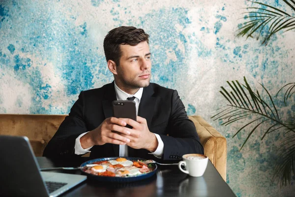 Üzletember ül a café segítségével a laptop számítógép van egy reggeli vagy vacsora eszik, mobiltelefon használata. — Stock Fotó