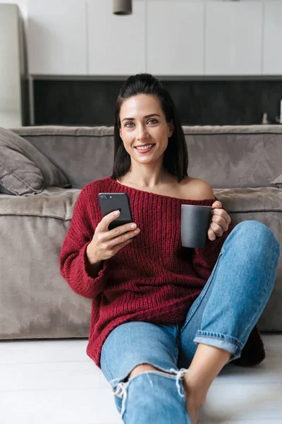 Όμορφη γυναίκα χρησιμοποιώντας κινητό τηλέφωνο πίνοντας καφέ σε κλειστούς χώρους στο σπίτι. — Φωτογραφία Αρχείου