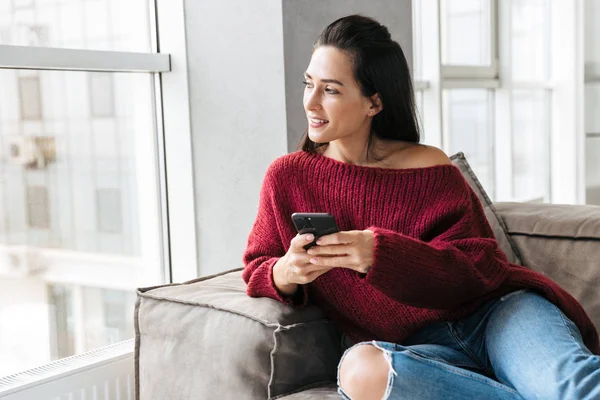 Schöne Frau drinnen im Haus auf dem Sofa mit dem Handy. — Stockfoto