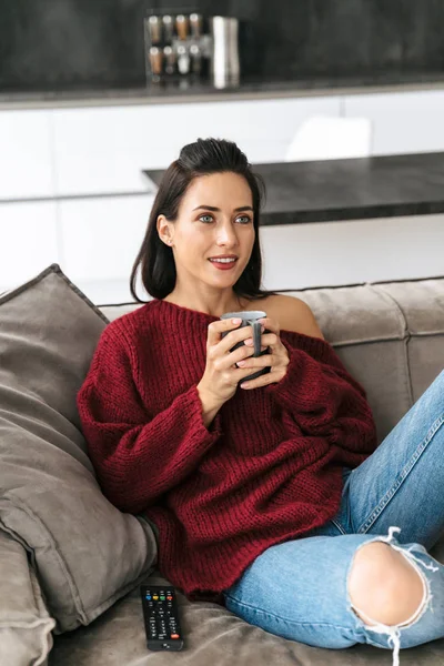 室内の女性家のソファの上でコーヒーを飲みながらテレビを見てください。. — ストック写真