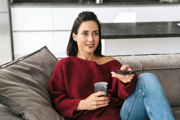 Nő beltéri otthon a kanapén tévénézés kávéfogyasztás. — Stock Fotó