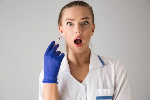 Beautiful young woman cosmetologist doctor isolated over grey wall background holding injection syringe. — Stock Photo, Image