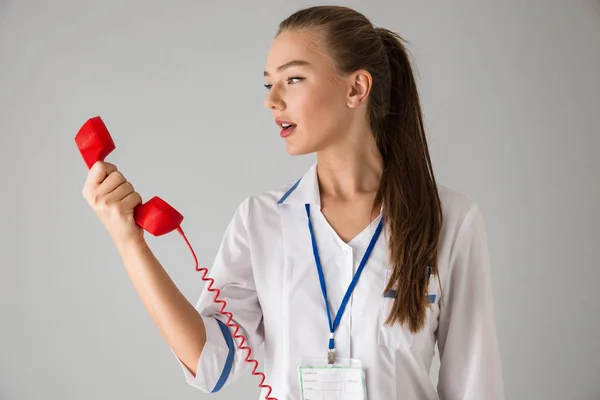 Beautiful young woman cosmetologist doctor isolated over grey wall background talking by telephone. — Stock Photo, Image