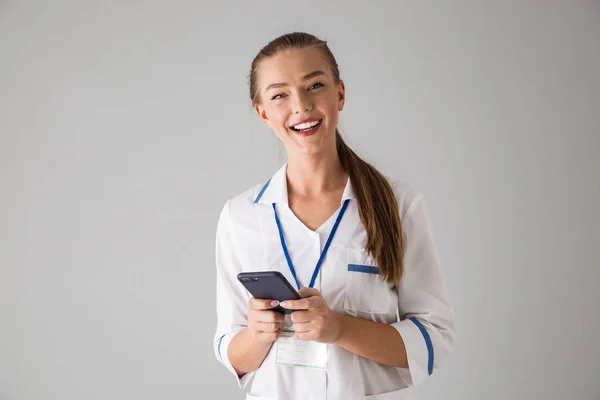 Szczęśliwa młoda kobieta lekarz kosmetolog na białym tle szare ściany trzymając telefon komórkowy za pomocą. — Zdjęcie stockowe