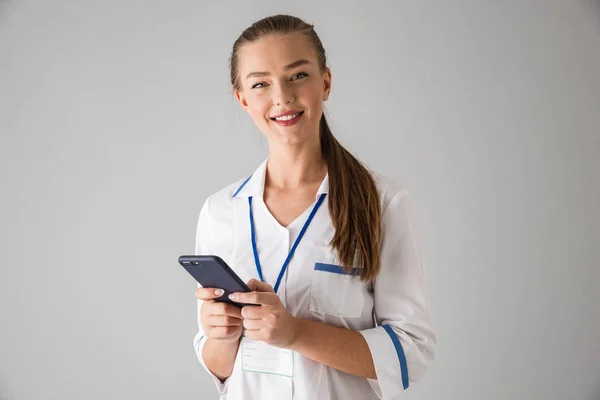 Счастливая молодая женщина косметолог врач изолирован на сером фоне стены держа мобильный телефон с помощью . — стоковое фото