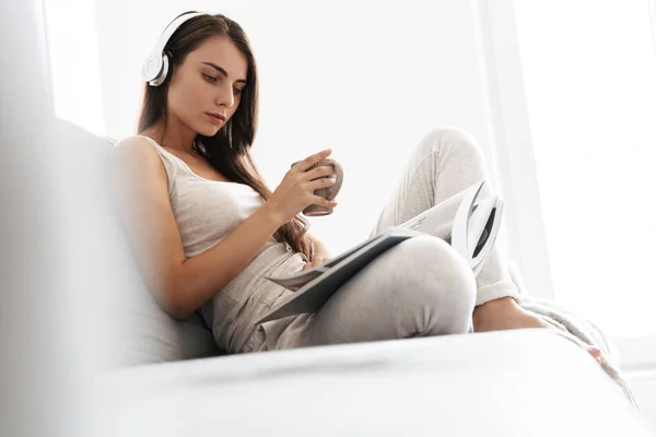 Giovane donna seduta sul divano al chiuso a casa ad ascoltare musica con le cuffie bere tè lettura rivista . — Foto Stock