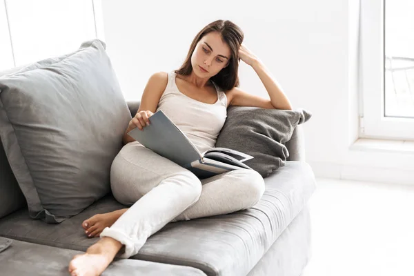 Beautiful young woman sitting on sofa indoors at home reading book. — Stock Photo, Image