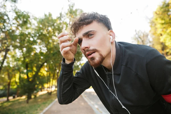 Schöner junger Sportler, der draußen im Park läuft und mit Kopfhörern Musik hört. — Stockfoto