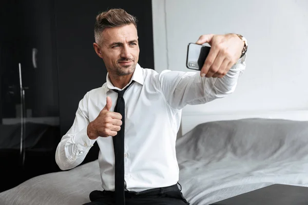 Bonito homem de negócios no quarto dentro de casa sentado na cama usando telefone celular tirar uma selfie . — Fotografia de Stock