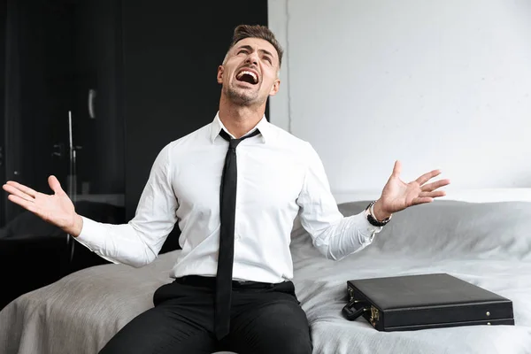 屋内で自宅の寝室で叫んで感情的なビジネスの男性がベッドの上に座ってください。. — ストック写真
