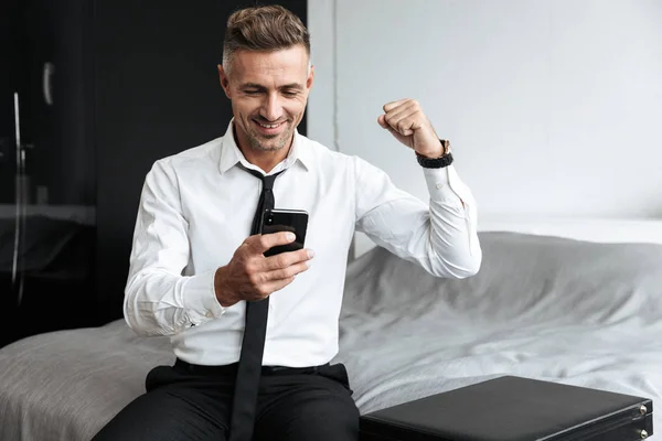 Business man in bedroom indoors at home sitting on bed using mobile phone make winner gesture. — Stock Photo, Image