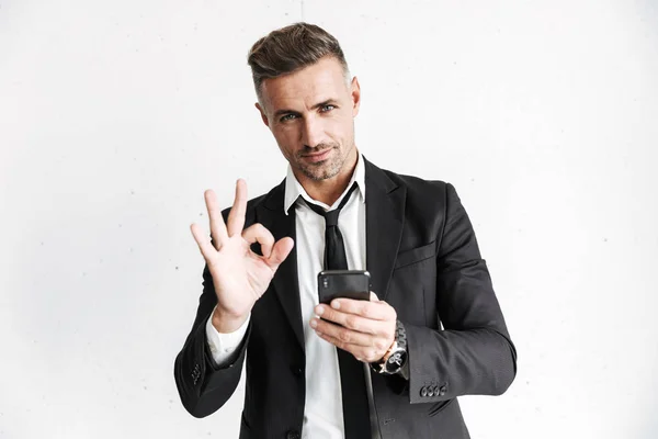 Hombre de negocios posando aislado sobre fondo de pared blanca utilizando el teléfono móvil que muestra gesto bien . — Foto de Stock