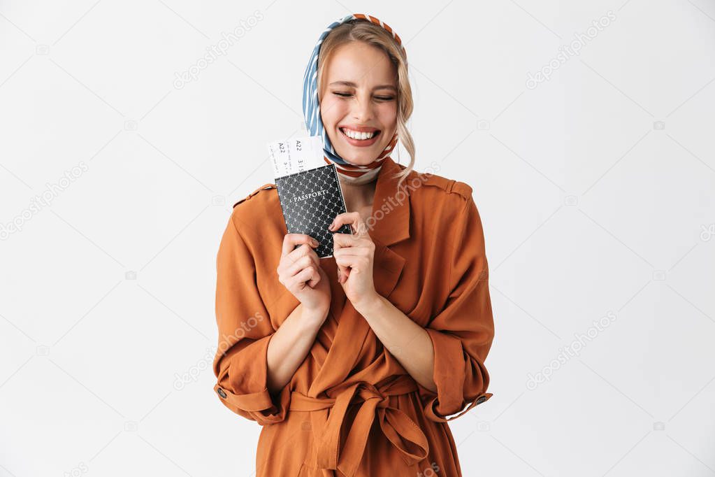 Beautiful excited young blonde pretty woman wearing silk scarf posing isolated over white wall background holding passport with tickets.